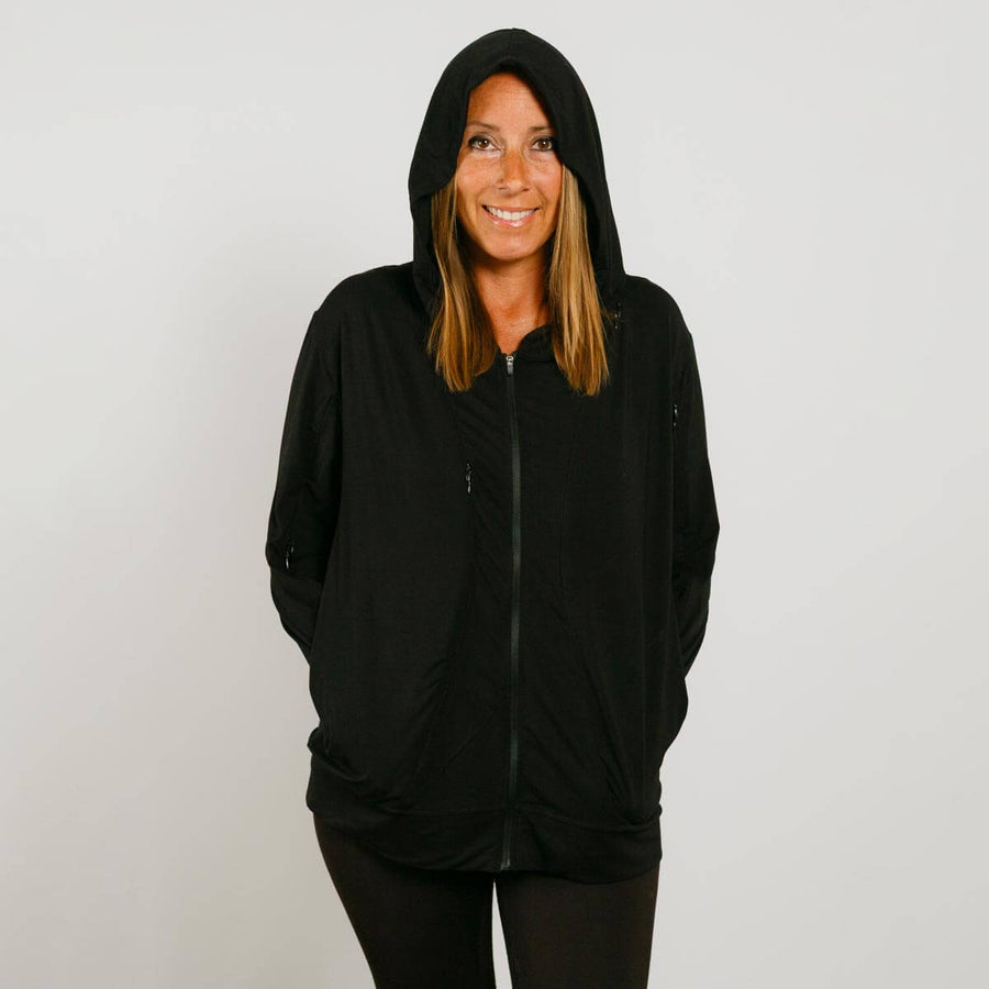 Woman wearing a black zip hoodie with hood up, hands in pockets, smiling at camera.