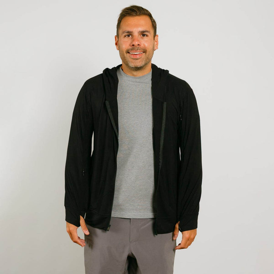Man wearing black zip hoodie over gray t-shirt and gray pants, smiling at camera.
