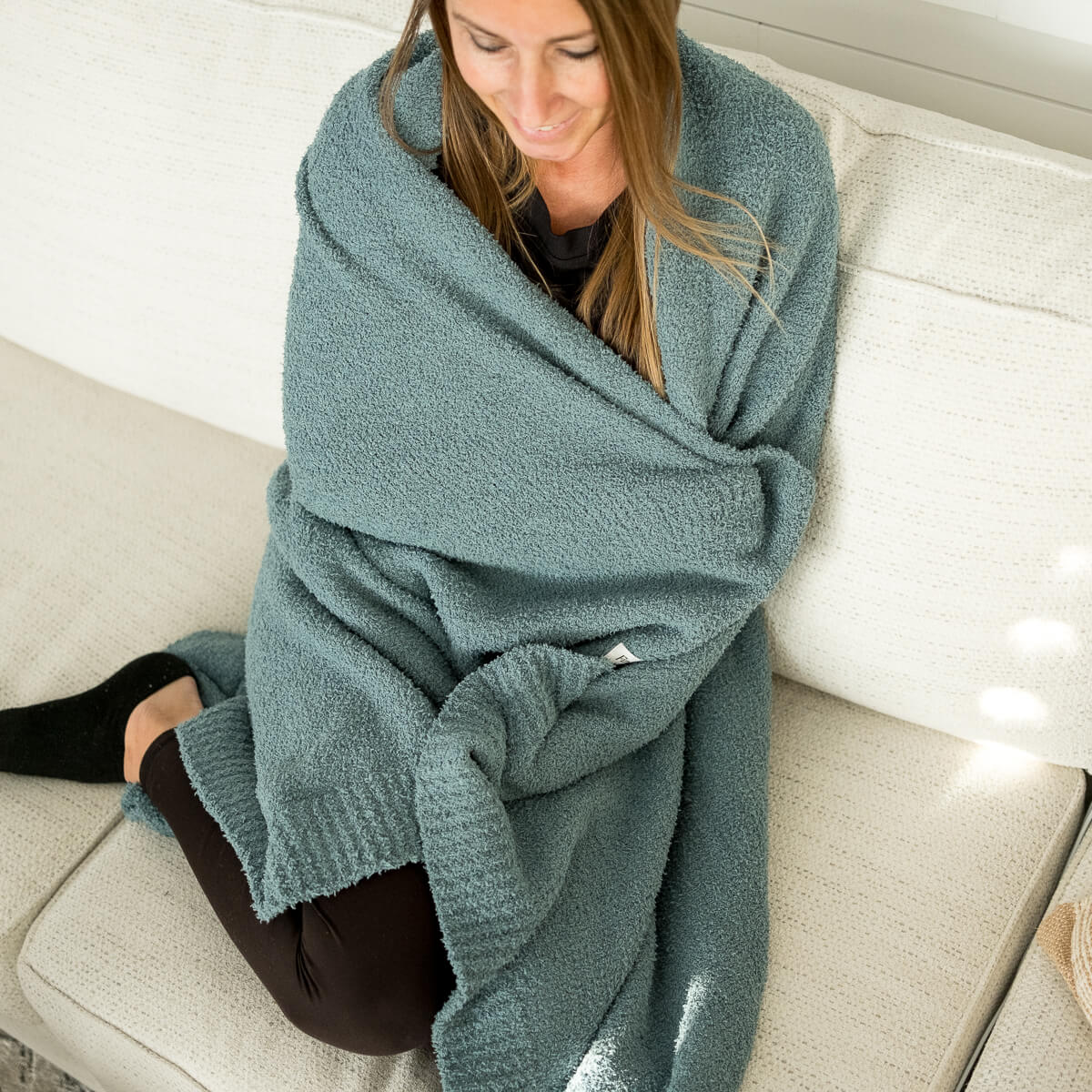A woman wrapped in a teal-blue plush blanket sits on a white couch.