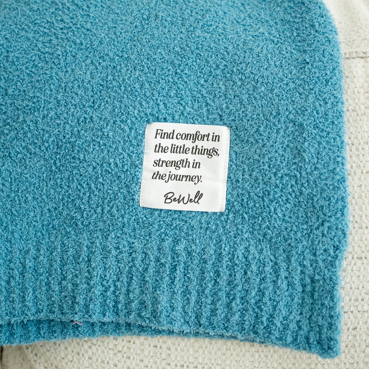 Close-up of a blue textured blanket with a white label that reads, "Find comfort in the little things, strength in the journey. Be Well."
