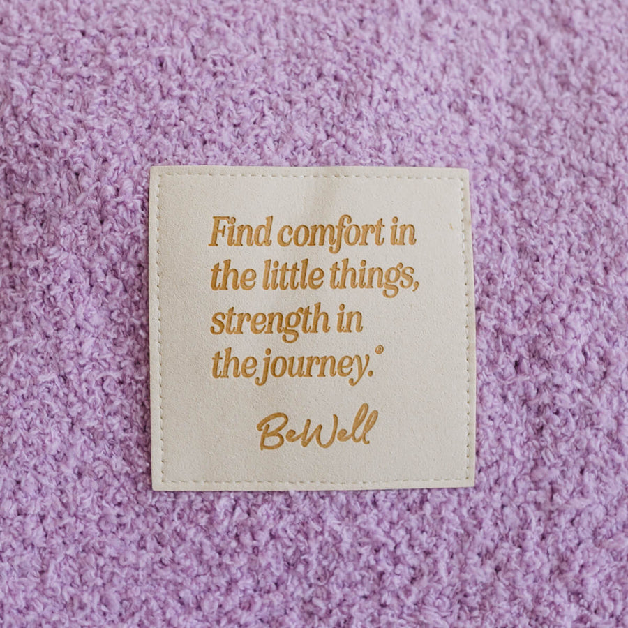 Close-up of cream infusion day blanket with gold inspirational quote detail on purple textured surface.