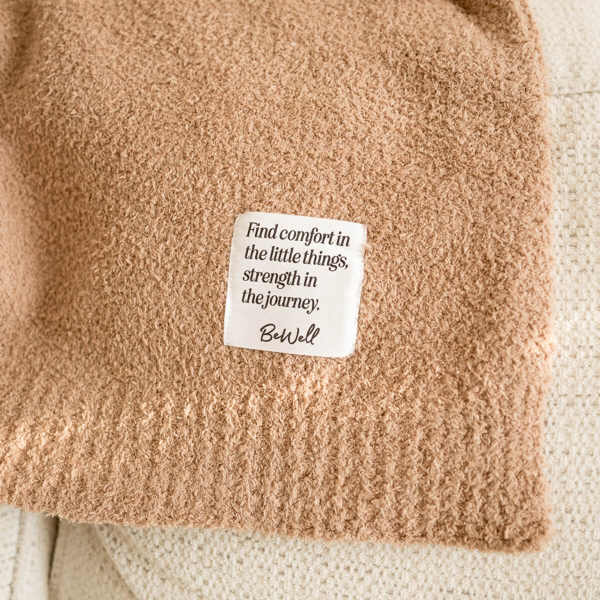 Beige, plush knit blanket with a "BeWell" tag.