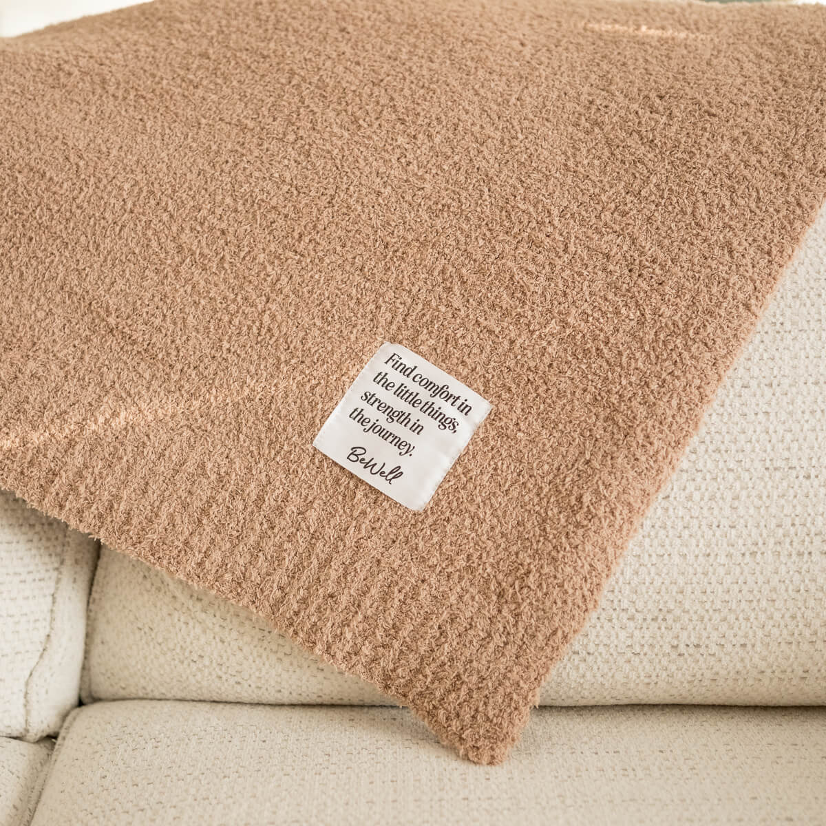 Beige fuzzy blanket with a white tag that reads "Find comfort in the little things, strength in the journey. BeWell".