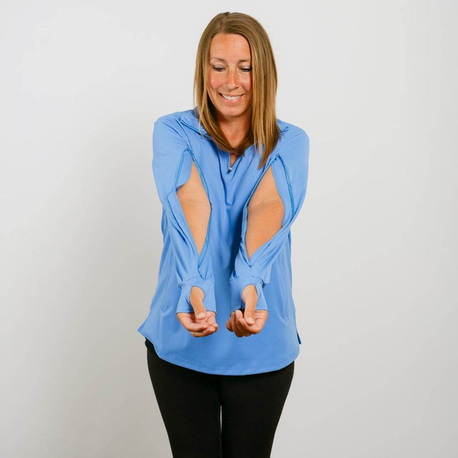 Woman wearing a light blue quarter zip top with elbow cutouts and black pants, smiling with hands outstretched.