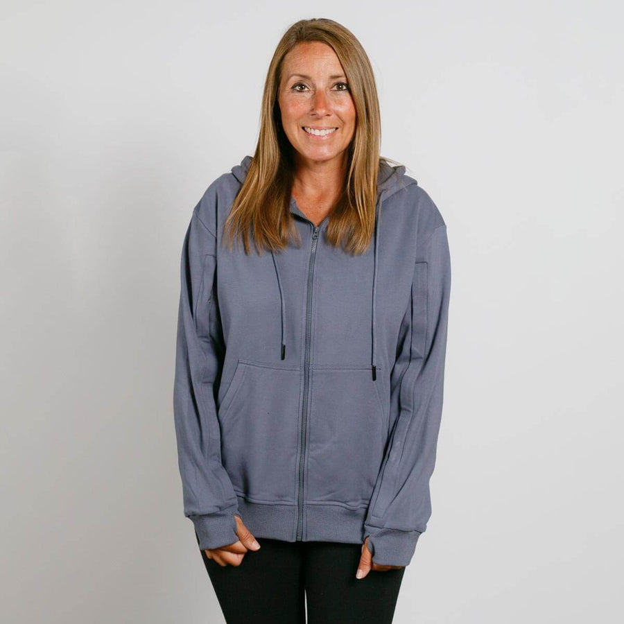 Woman wearing a slate blue zip-up hoodie over black pants, smiling at camera against white background.