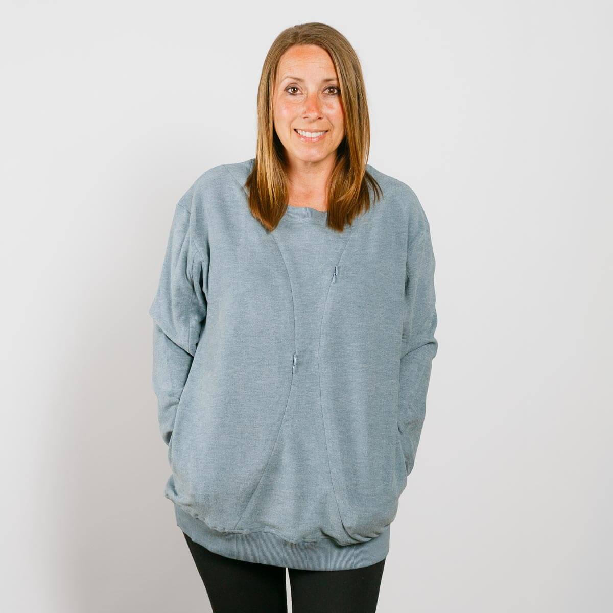 Woman wearing a light blue crewneck sweatshirt with hands in pockets, smiling at camera.