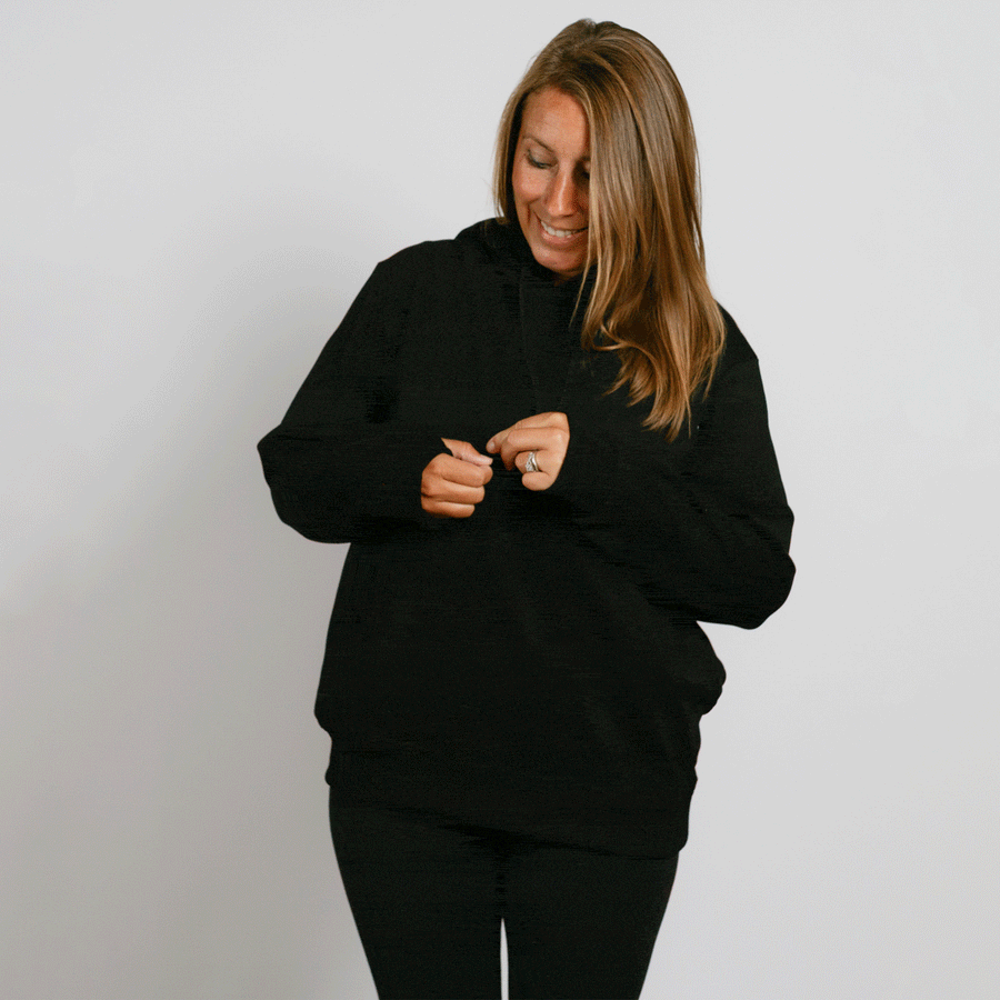 Woman wearing black hoodie with kangaroo pocket, looking down with hands near chest.