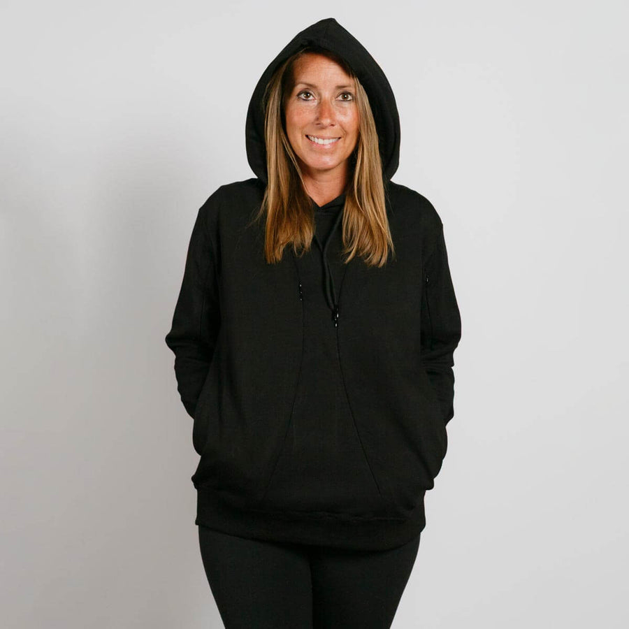 Woman wearing a black hoodie with hands in pockets, smiling at camera against white background.