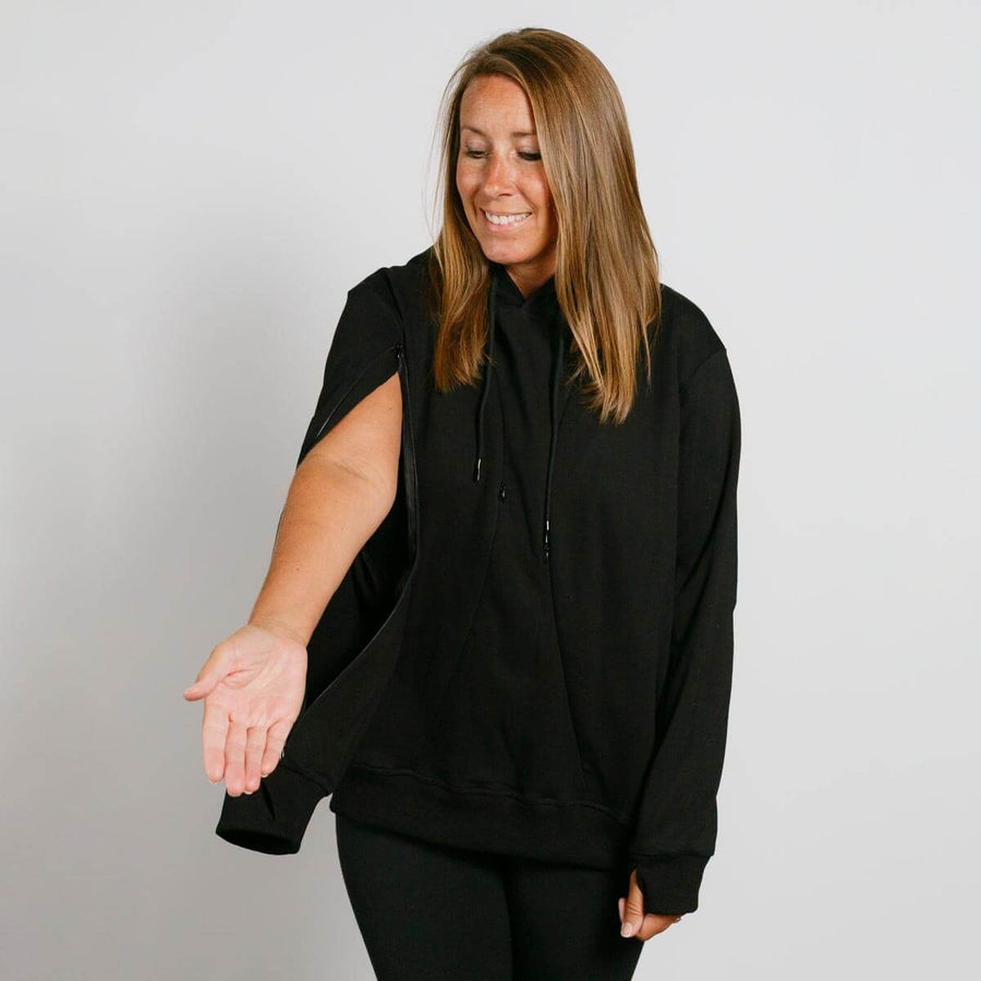 Woman wearing a black sleeveless hoodie with short sleeves over black pants, smiling downward.