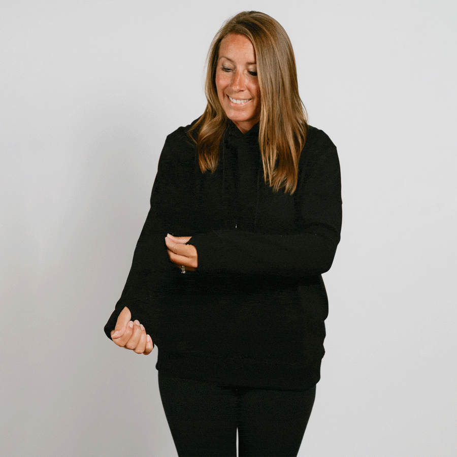 Woman wearing a black hoodie and black pants, smiling while looking downward against a white background.