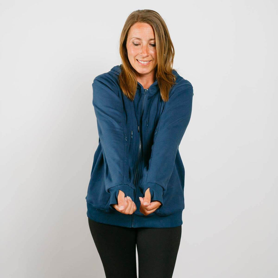 Woman wearing a dark blue zip hoodie and black pants, smiling with hands clasped together.