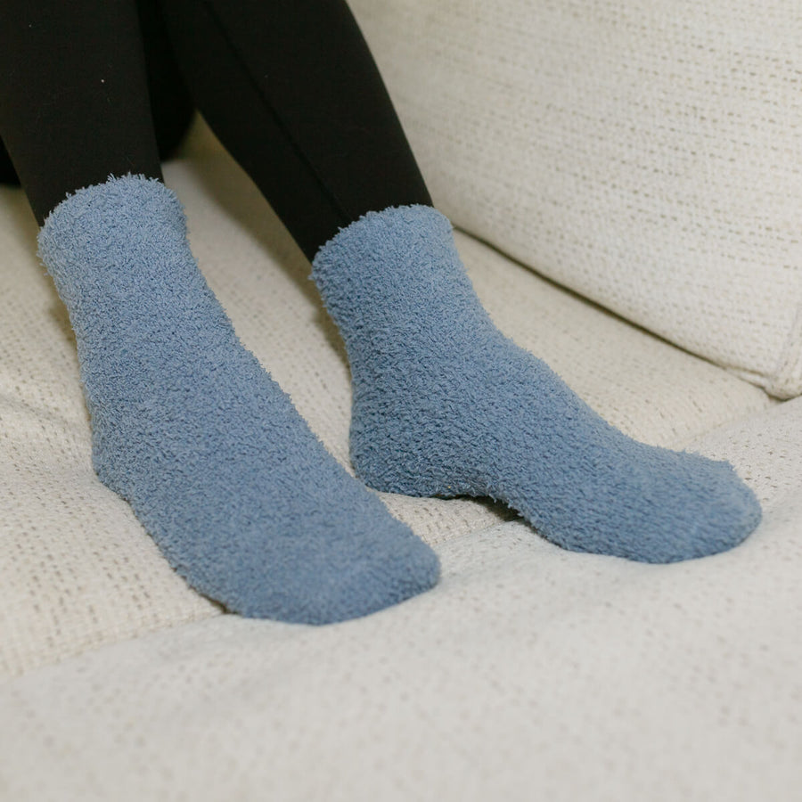 Person wearing blue cozy rest day socks with black pants, seated on cream-colored furniture.