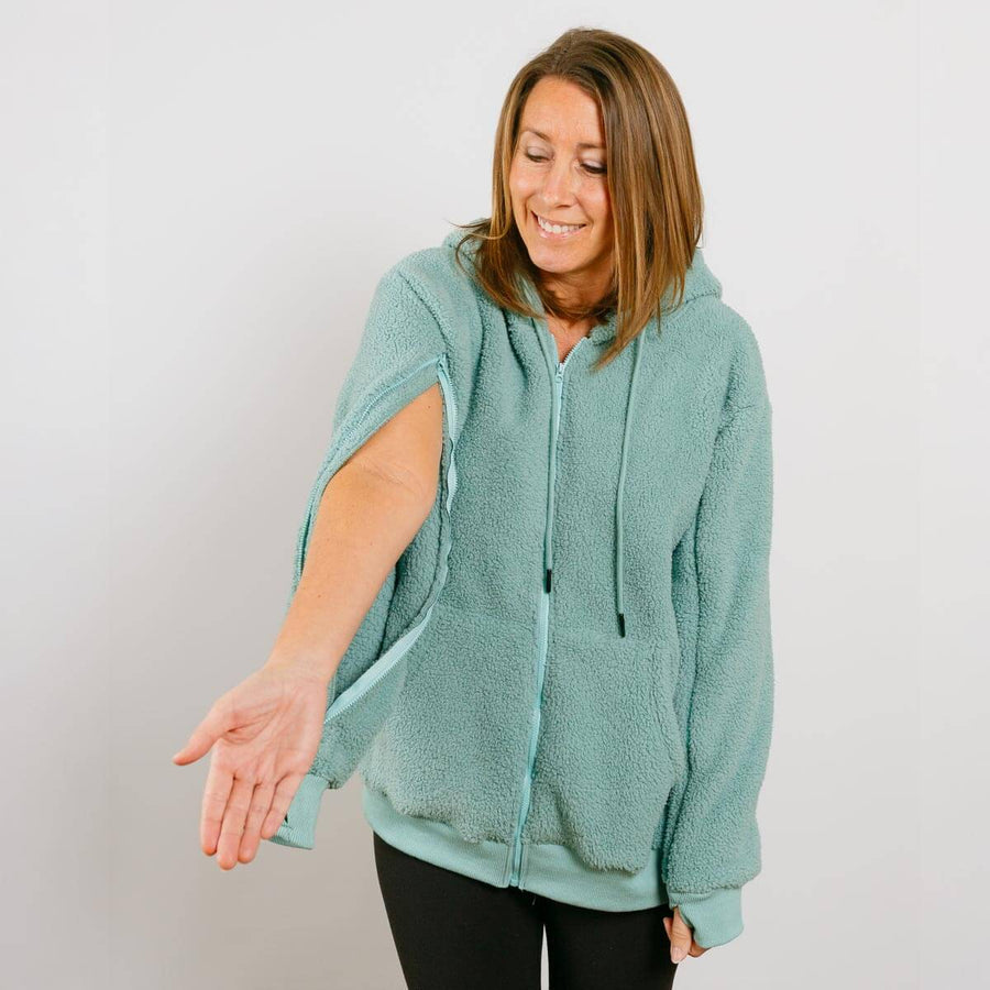 Woman wearing sage green sherpa zip hoodie with left sleeve access opening visible.