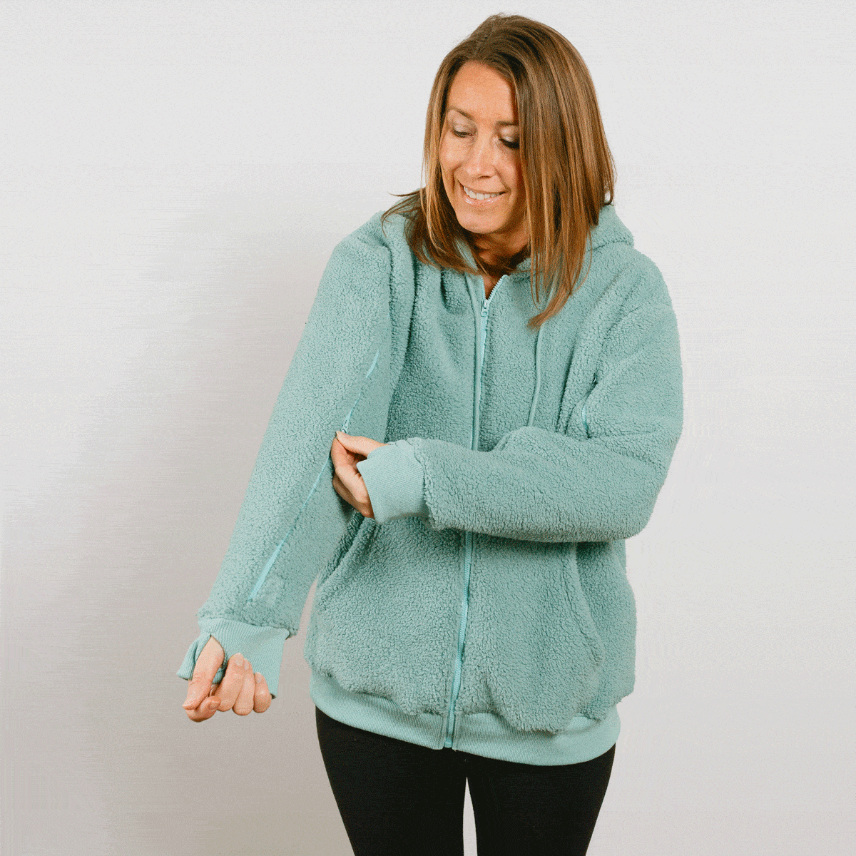 Woman wearing sage green sherpa zip hoodie with front zipper, smiling while adjusting sleeve.