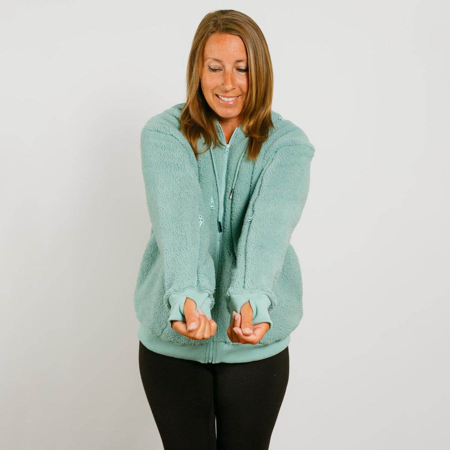 Woman wearing a seafoam color zip hoodie with thumb holes over black pants, smiling while looking down.