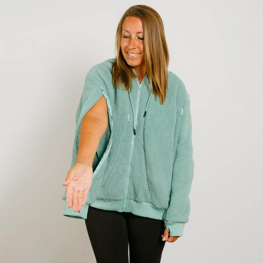 Woman wearing a seafoam color sherpa zip hoodie with black pants, smiling while looking down.