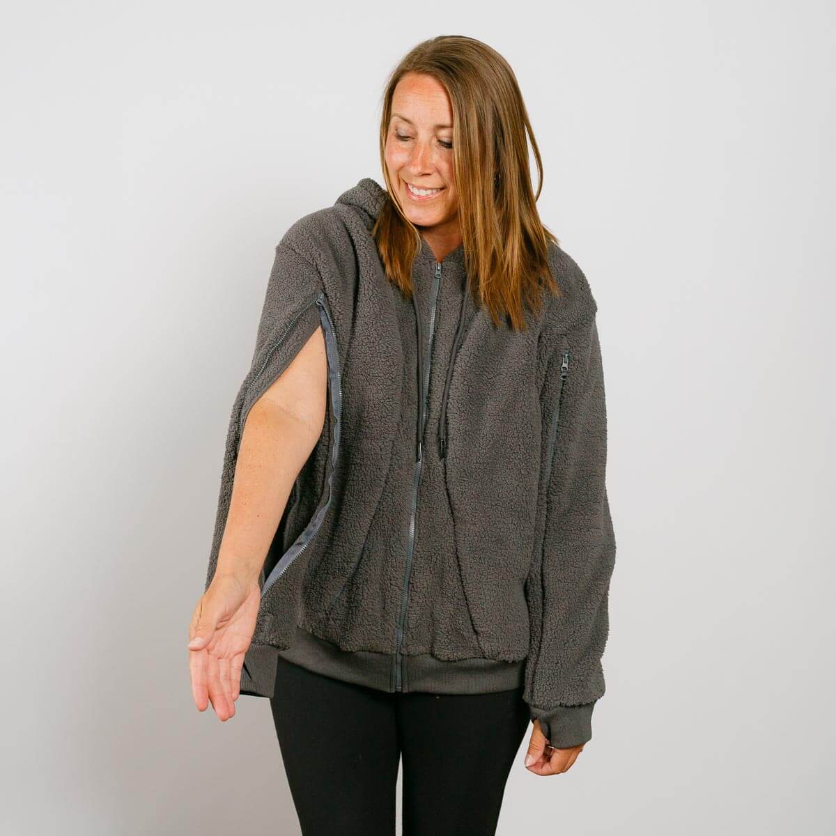 Woman wearing a gray sherpa zip hoodie with cutout sleeves and black pants, smiling downward.