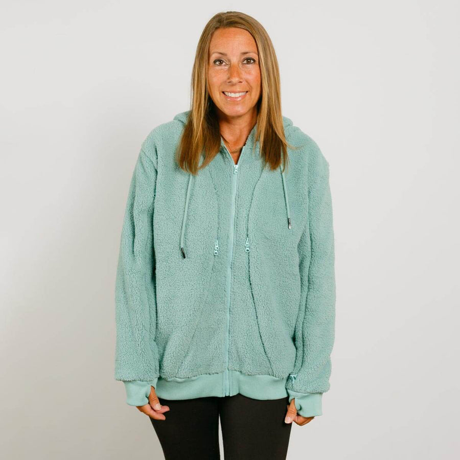 Woman wearing a seafoam color sherpa zip-up hoodie with drawstrings over black pants.