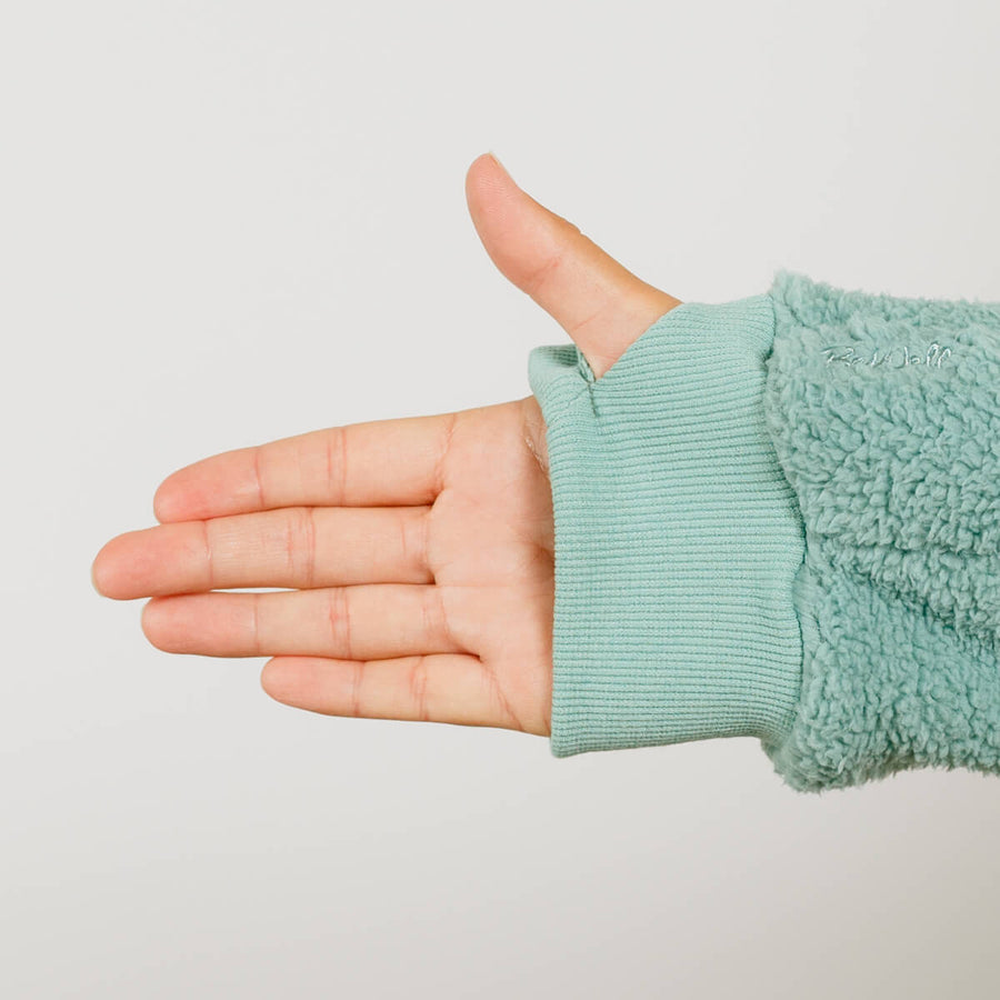 Womans's hand extended from seafoam color fuzzy sherpa sleeve with ribbed cuff against neutral background.