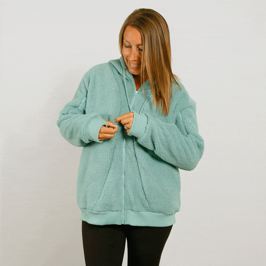 Woman wearing a seafoam color sherpa zip hoodie over black pants, zipping the front closed.