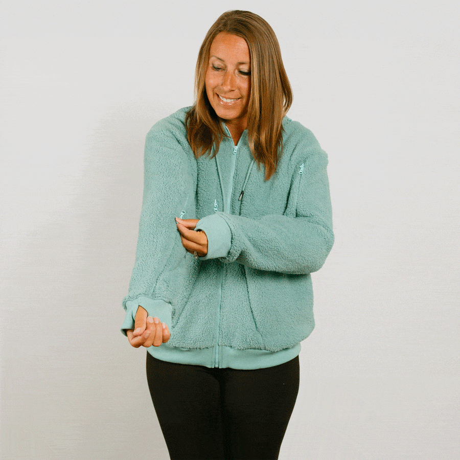 Woman wearing a seafoam color zip-up hoodie with black pants, smiling while looking down.