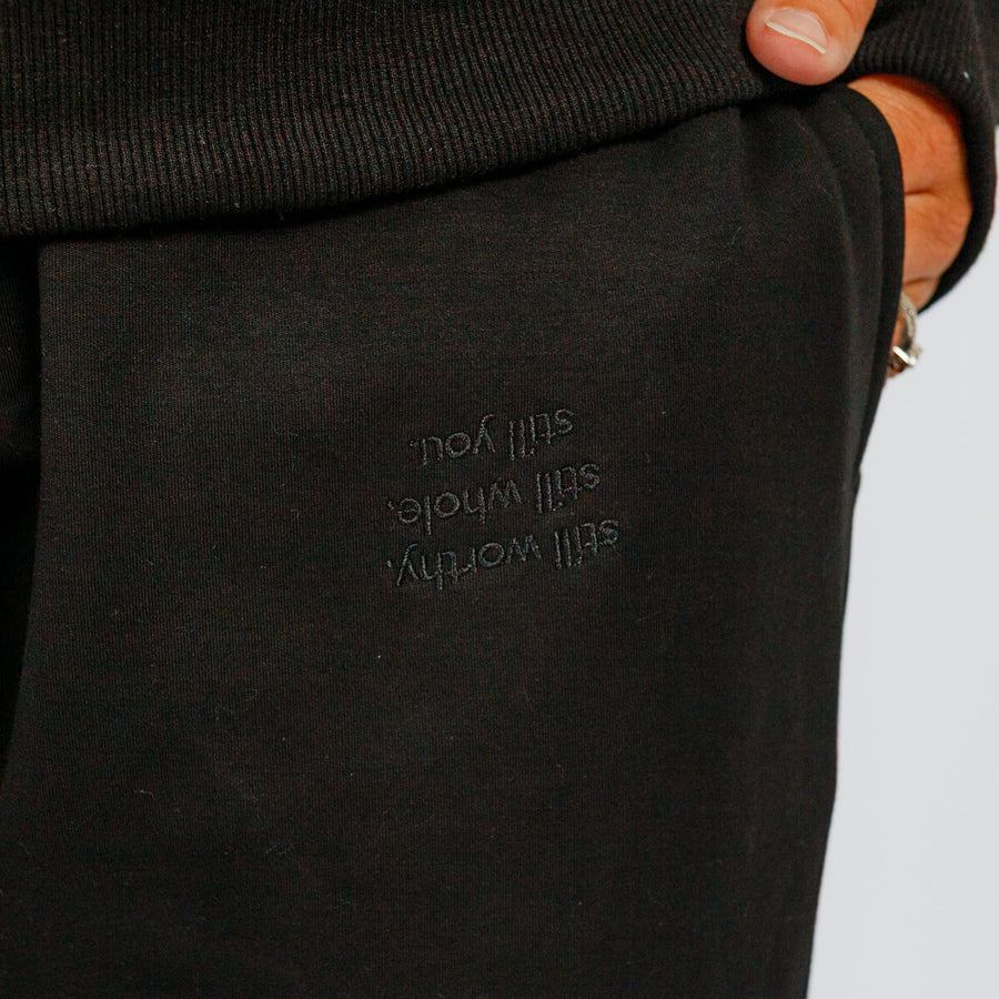 Black sweatpants with embroidered text on the leg, worn with a dark sweater and orange-toned shoes.
