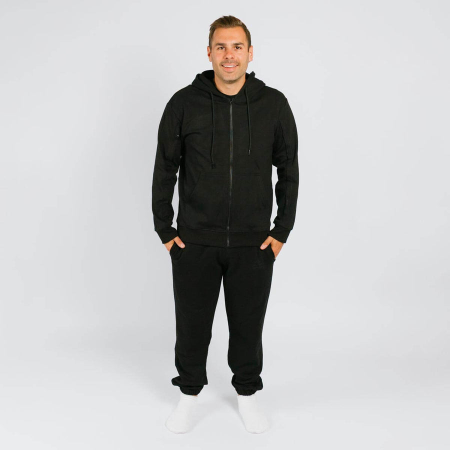 Man wearing black zip-up hoodie and black sweatpants with white socks against gray background.