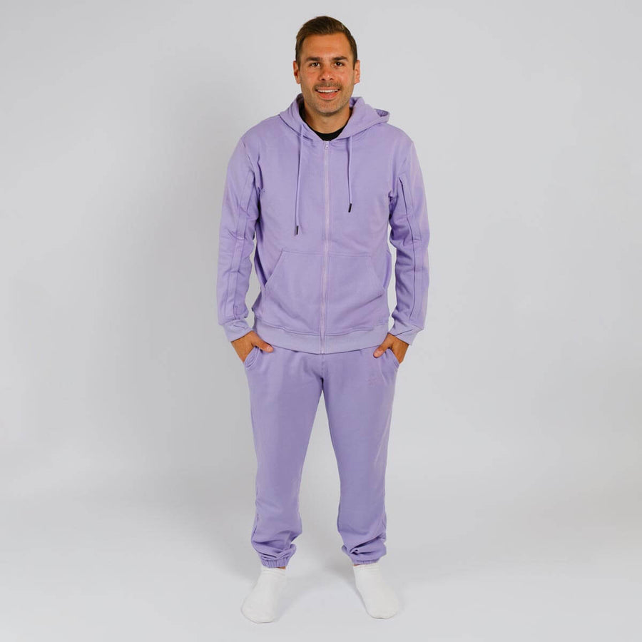 Man wearing lavender zip-up hoodie and matching sweatpants standing against white background.