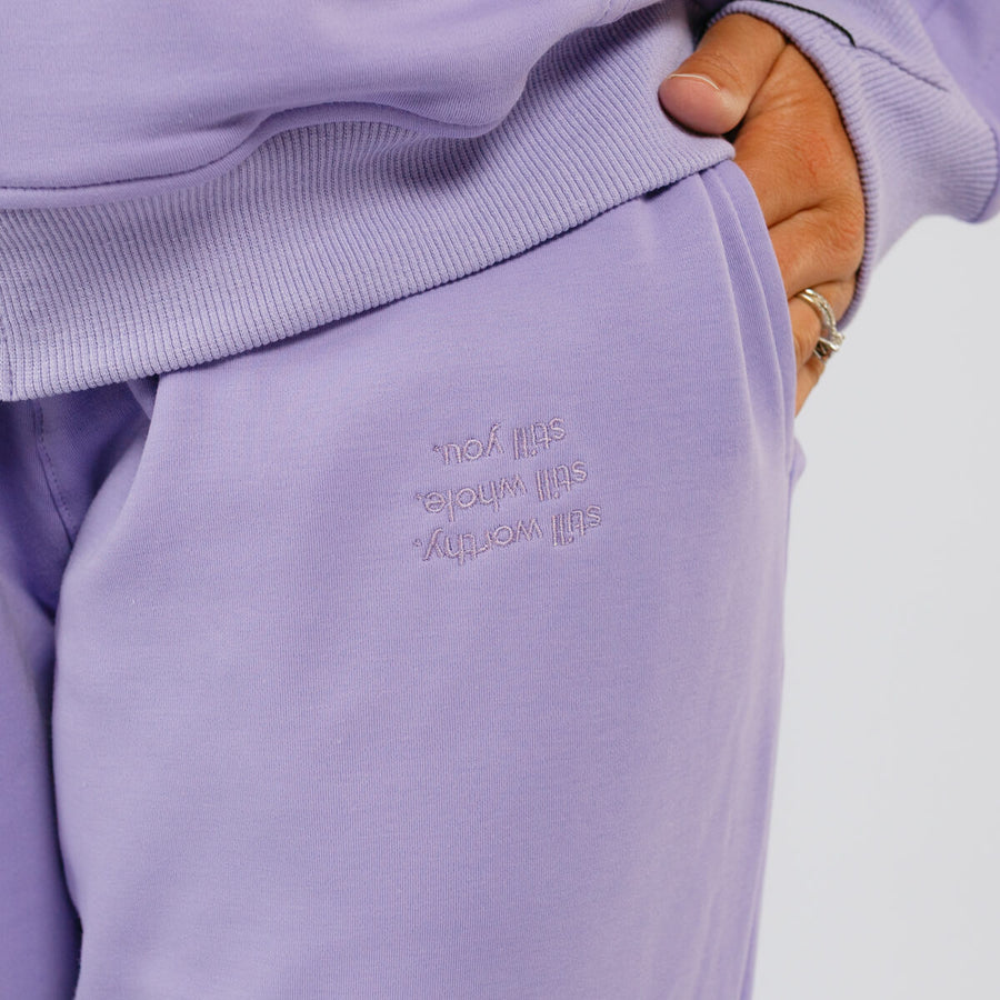 Purple sweatpants with embroidered text detail on the thigh, shown on a person wearing a lavender hoodie.