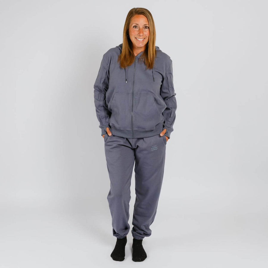 Woman wearing a gray zip-up hoodie and matching slate blue sweatpants with black socks against a white background.