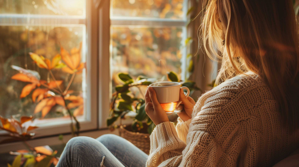 Woman in cozy sweater holding warm cup by window, finding peaceful moment during autumn day.