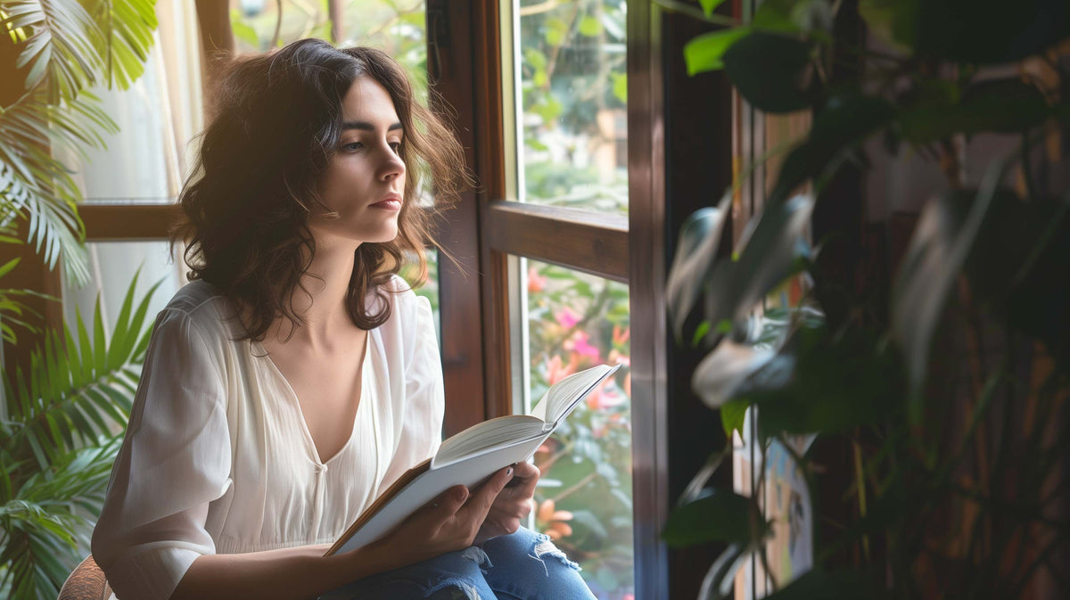 Woman reading by a window surrounded by plants, reflecting on managing chronic illness emotions.