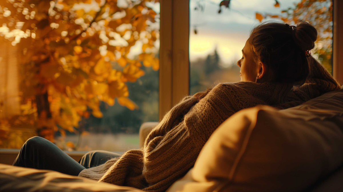 Person in soft sweater relaxing by window on autumn day, gazing at golden fall landscape.