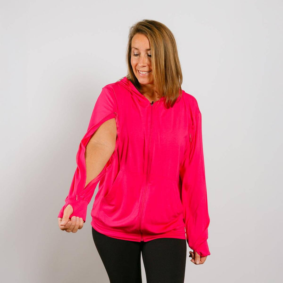 Woman wearing a bright pink zip-up hoodie with cutout sleeves over black pants, smiling downward. #NEW Woman wearing a bright pink hoodie with cut-out sleeves on a plain background