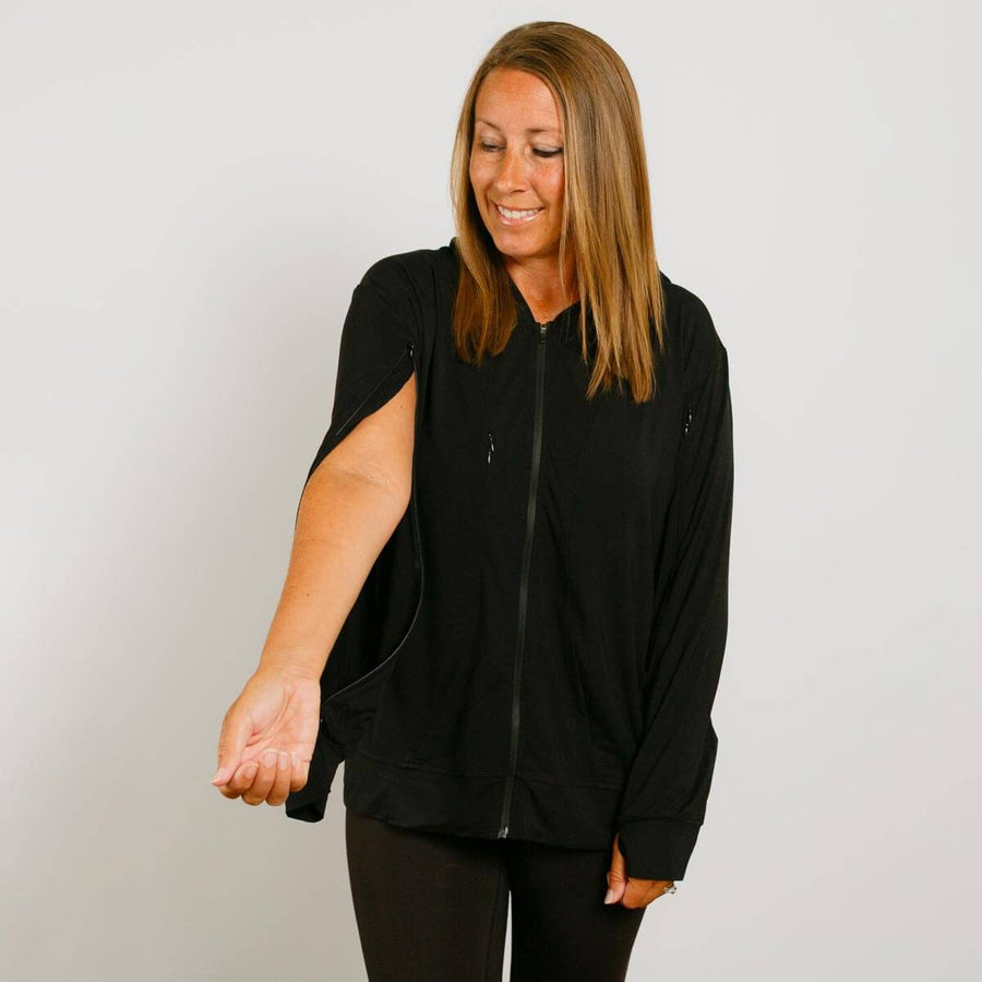 Woman wearing a black zip-up hoodie with short sleeves over dark pants, smiling downward.