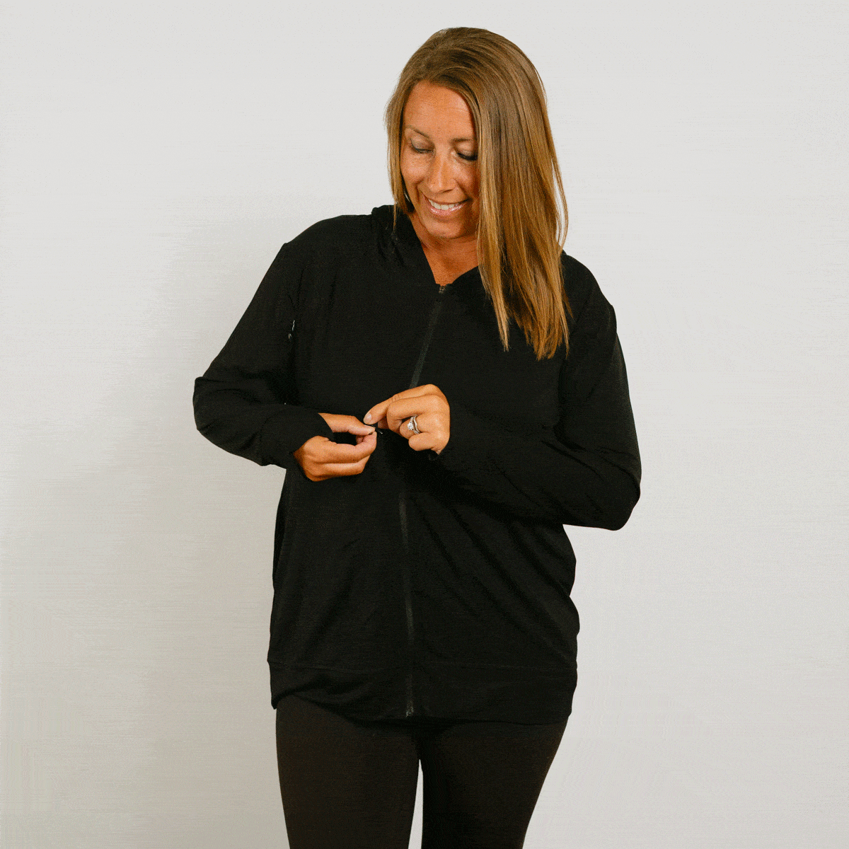 Woman wearing a black zip hoodie, smiling while zipping the front closure. #gif
