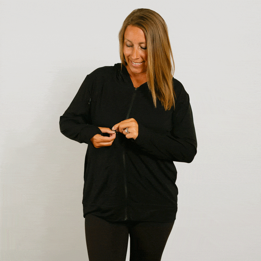 Woman wearing a black zip hoodie, smiling while zipping the front closure.