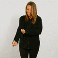 Woman wearing black zip hoodie with hands relaxed at sides, smiling downward against white background.