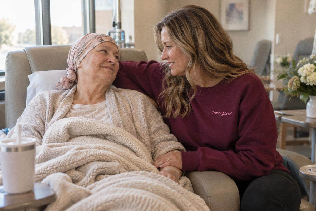 Woman in the plum Care Giver crewneck with "care giver" embroidered on the chest, smiling at an older woman in a head scarf resting in a treatment chair.