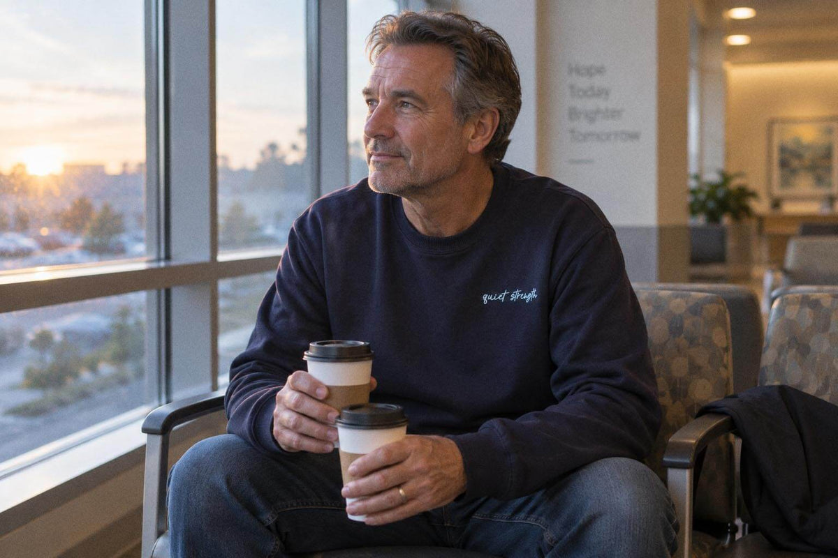 Man in the navy Quiet Strength crewneck holding two coffees, looking out a hospital waiting room window at sunrise.