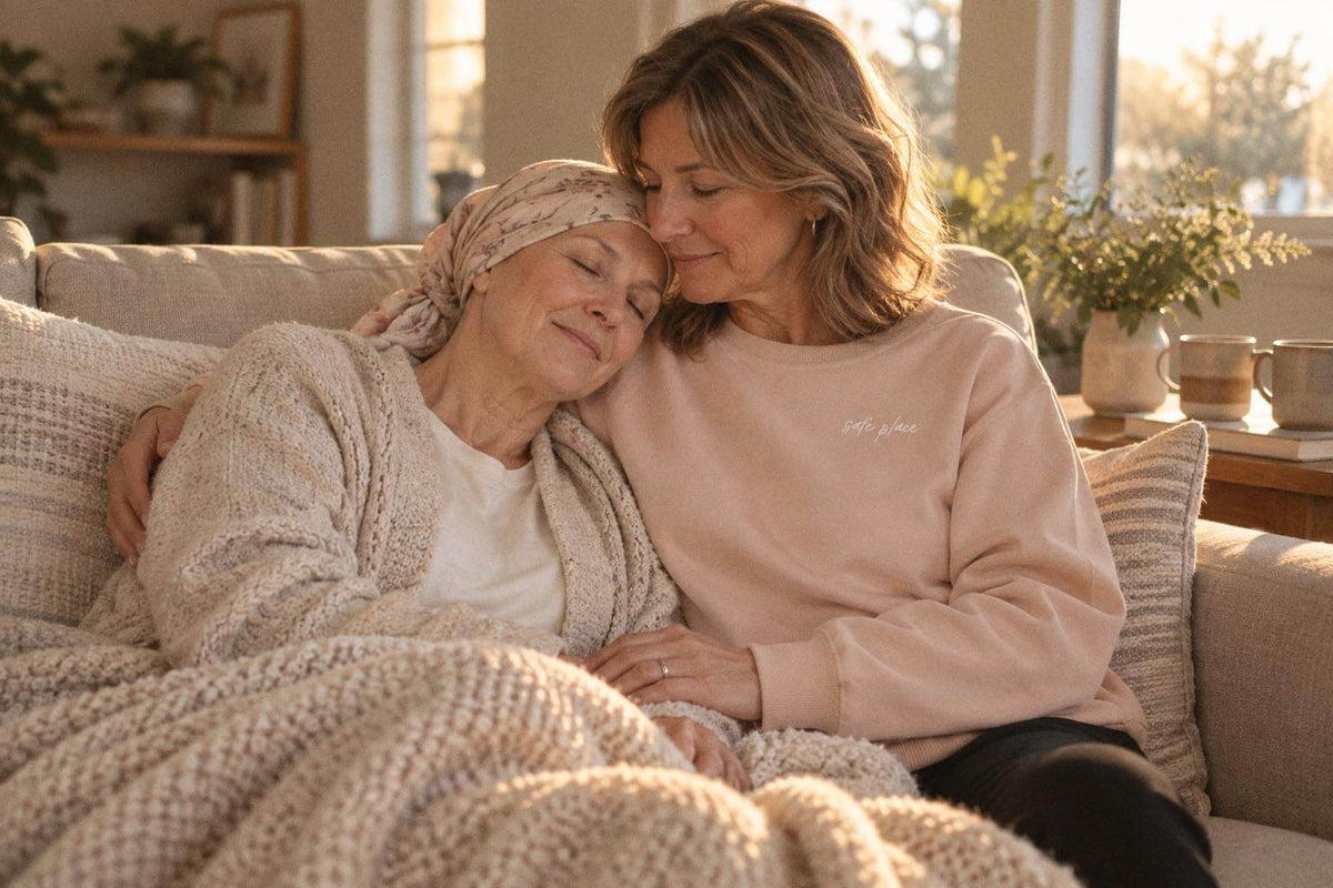 Woman in the beige Safe Place crewneck on a couch at home, holding an older woman in a head scarf who rests her head on her shoulder.