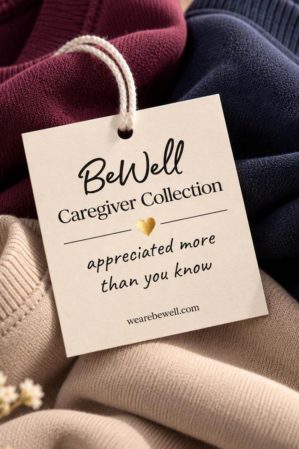 The cream BeWell Caregiver Collection tag with a gold foil heart, reading "appreciated more than you know," resting on the plum, navy, and beige crewnecks.