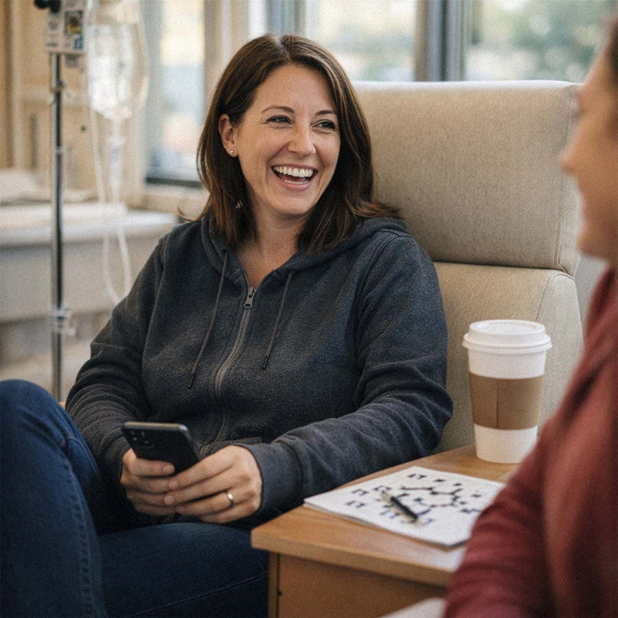 Woman in dark hoodie smiling while holding phone, sitting in medical chair with coffee cup nearby.