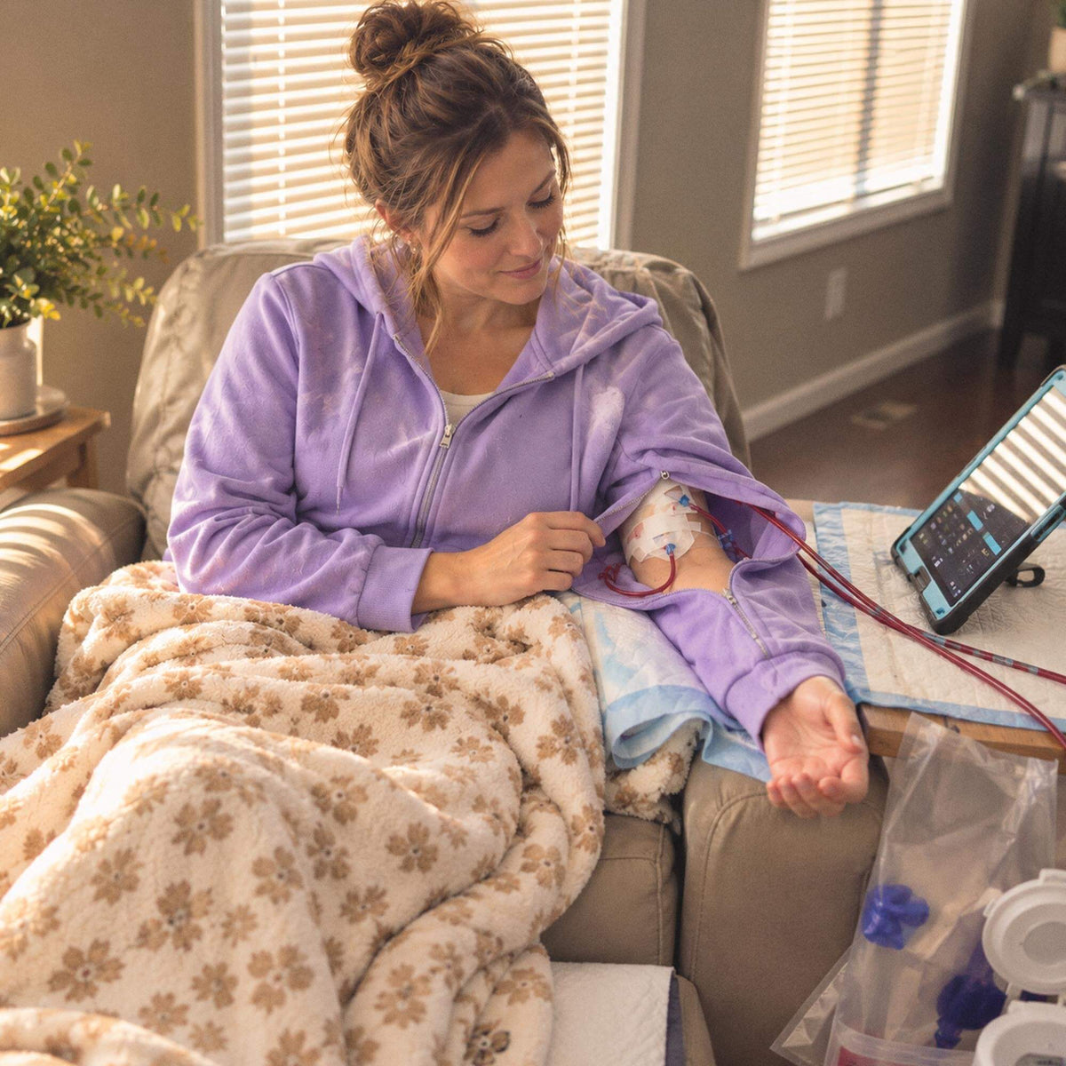 Woman in a lavender BeWell IV Zip Hoodie relaxing in a treatment recliner under a cozy blanket, the sleeve unzipped around her treatment lines so her arm stays covered.