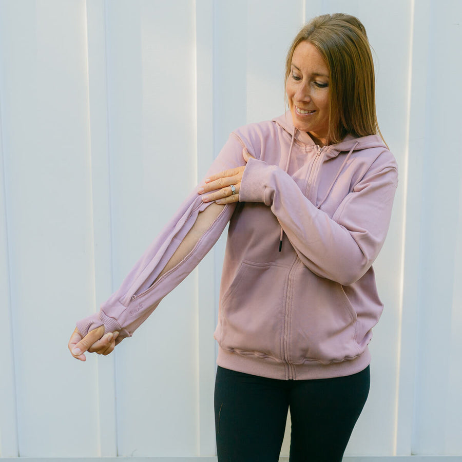Woman wearing a pink hoodie adjusting her arm in front of a white wall. #RESTOCK