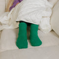 Person wearing green Rest Day socks while bundled under white quilted blanket on bed.