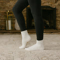 Person wearing black leggings and white crew socks standing indoors by fireplace.