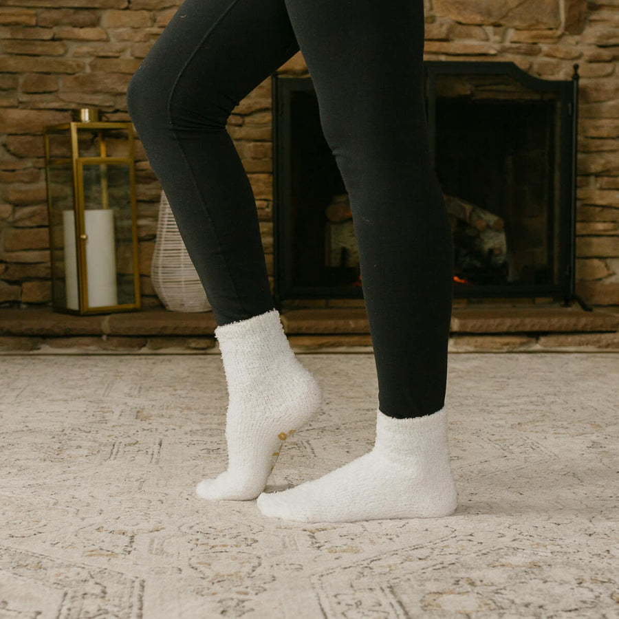 Person wearing black leggings and white crew socks standing indoors by fireplace.
