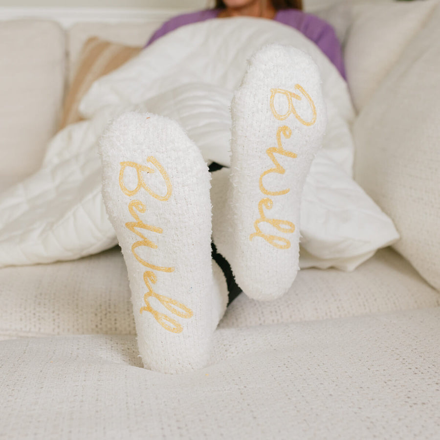 Woman wearing white Rest Day socks with gold text, feet up on couch.