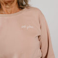 Woman wearing a beige crewneck sweatshirt with "safe place" embroidered in white script.